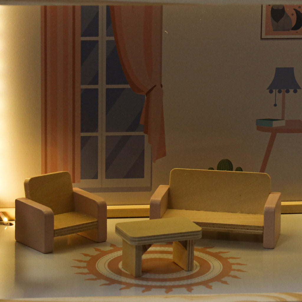 Klimatyczny pokój dzienny w drewnianym domku dla lalek, z różowymi akcentami. Widoczne miniaturowe drewniane mebelki: różowa sofa, fotel i stolik kawowy, efektownie oświetlone ciepłym światłem LED, idealne do magicznej zabawy.
