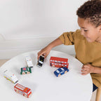 Chłopiec bawi się zestawem Drewnianych Samochodów Londyn: czerwony autobus piętrowy, czarna taksówka, wóz strażacki, furgonetka policji, karetka pogotowia, mini i auto sportowe. Te ekologiczne pojazdy dla dzieci z certyfikowanego drewna FSC rozwijają wyobraźnię.