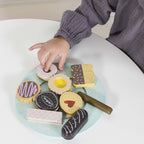 Dziecięca dłoń sięga po apetyczne, pastelowe drewniane ciasteczka i słodkości z Drewnianych ciasteczek zestaw na eleganckim talerzu, idealne do bezpiecznej, realistycznej zabawy.