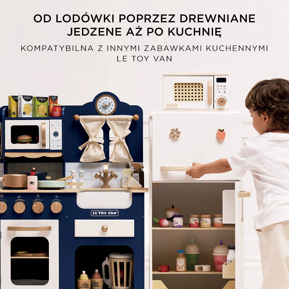 Drewniana kuchenka mikrofalowa Le Toy Van dla dzieci w białym kolorze z elementami drewna, widoczna w pełnej kuchni zabawkowej. Chłopiec otwiera obok niej lodówkę, szykując się do kreatywnej zabawy w gotowanie.