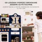 Drewniana kuchenka mikrofalowa Le Toy Van dla dzieci w białym kolorze z elementami drewna, widoczna w pełnej kuchni zabawkowej. Chłopiec otwiera obok niej lodówkę, szykując się do kreatywnej zabawy w gotowanie.