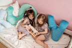 Dwie urocze dziewczynki czytające książkę w dziecięcym pokoju, otoczone pluszowymi poduszkami minky Dream Pillow. Miękkie poduszki z wypustkami tworzą przytulne miejsce do zabawy i relaksu.