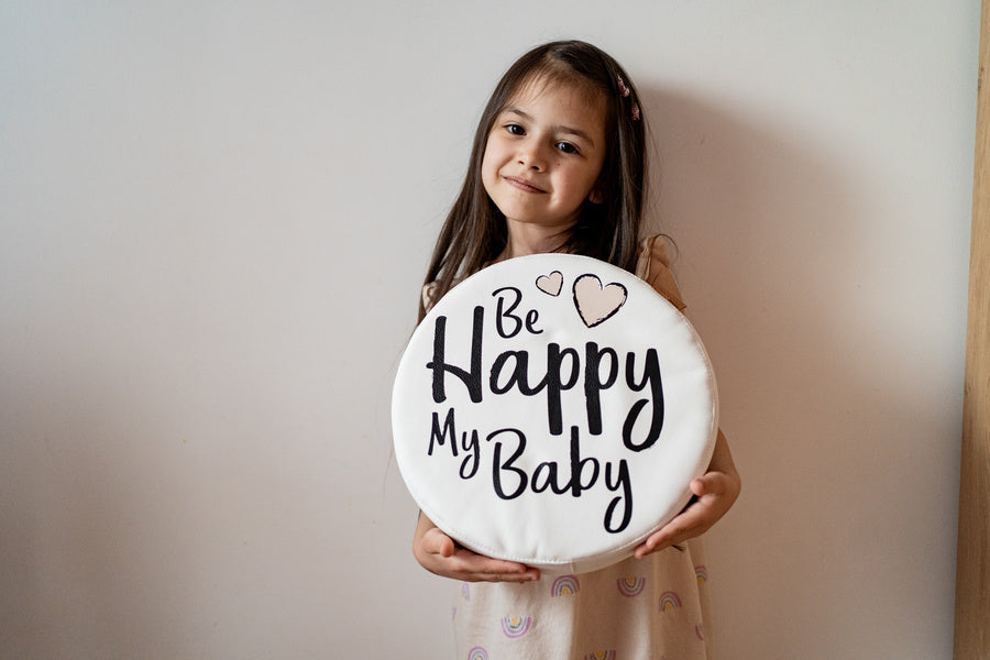 Uśmiechnięta dziewczynka prezentująca białą, okrągłą poduszkę z napisem "Be Happy My Baby". Idealna poduszka dekoracyjna z ekoskóry do pokoju dziecięcego, kącika zabaw, zapewniająca komfort i styl.