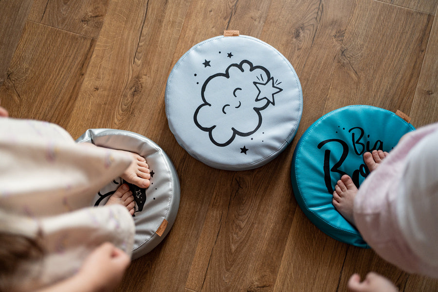 Zestaw 3 okrągłych poduszek z ekoskóry LITTLE PRINCESS o grubości 6 cm, z motywami chmurki i gwiazdki, na których dzieci bawią się na drewnianej podłodze. Idealne do pokoju dziewczynki.